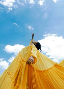 Woman in a flowing yellow dress posing under blue sky with clouds