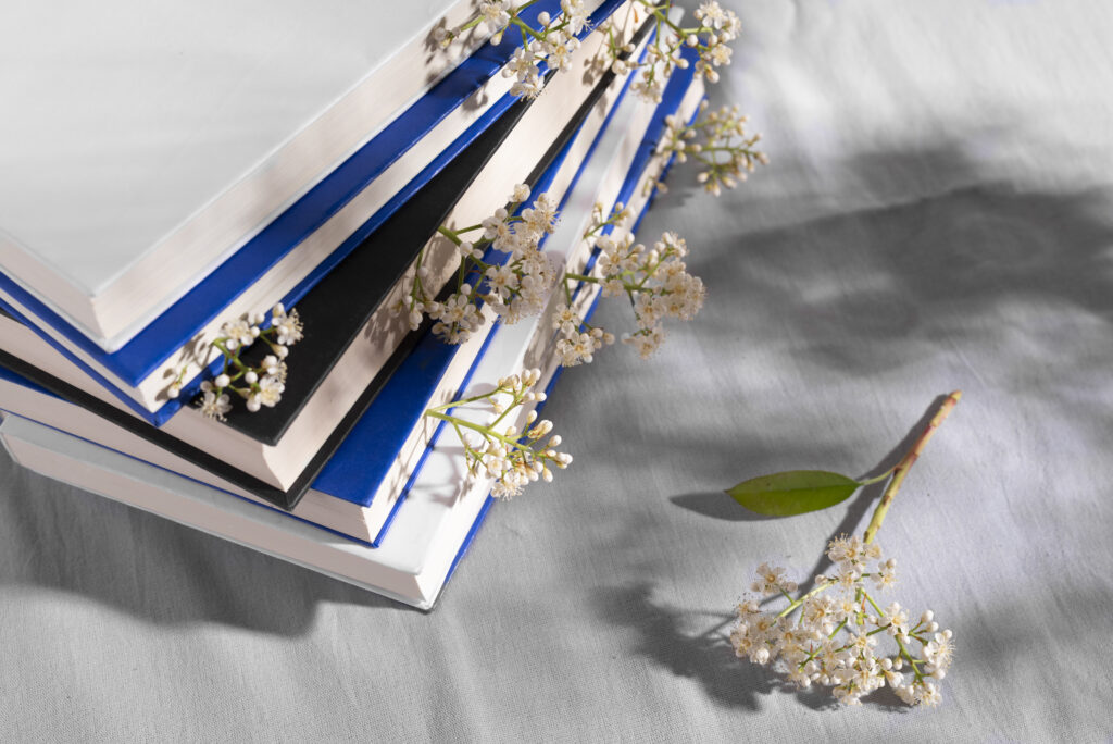 tack of books with blue and black covers, adorned with delicate white spring blossoms, placed on a soft gray fabric surface