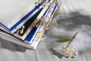 tack of books with blue and black covers, adorned with delicate white spring blossoms, placed on a soft gray fabric surface